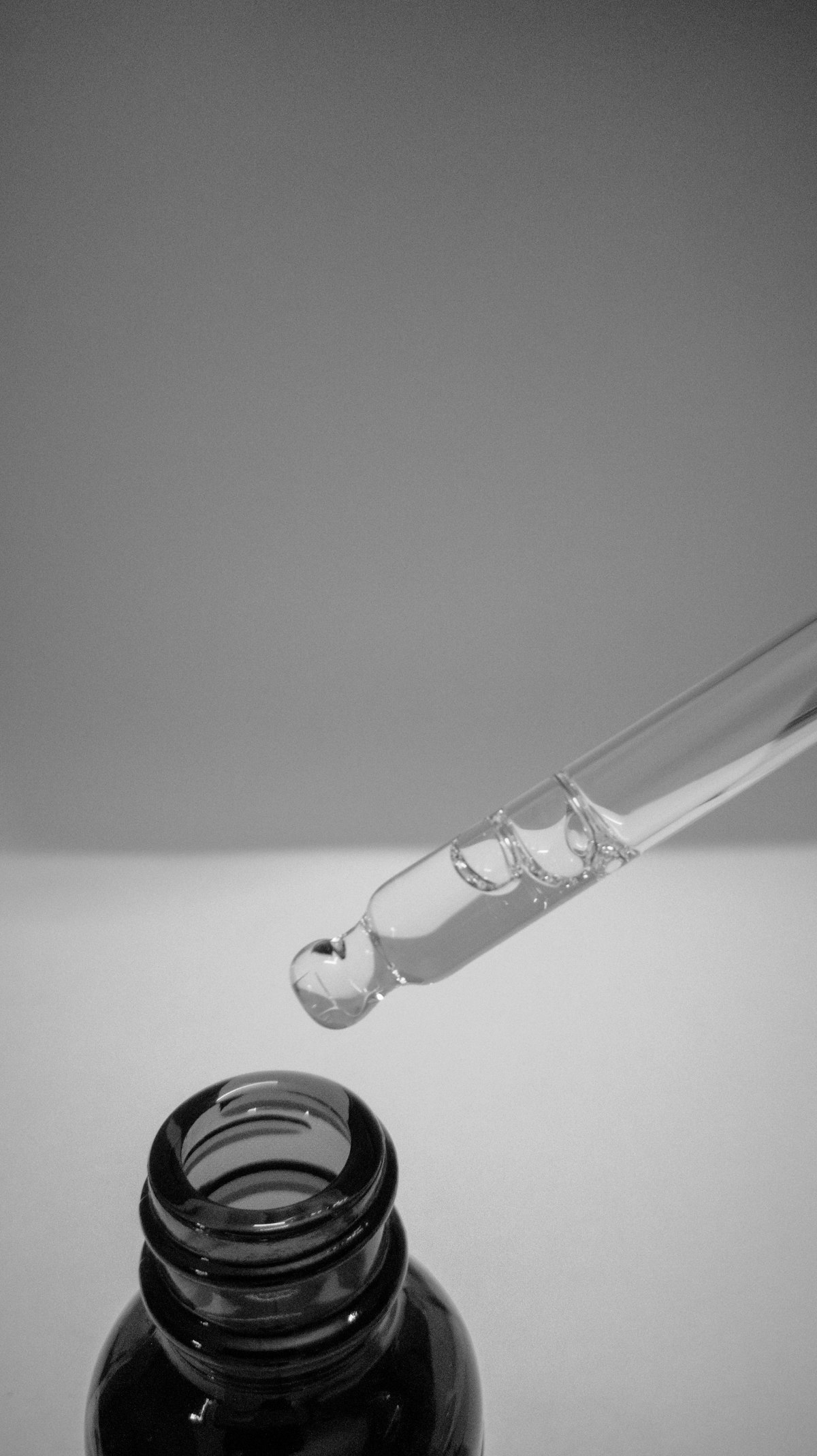 Glass pipette releasing a drop of oil into a dark bottle, in monochrome